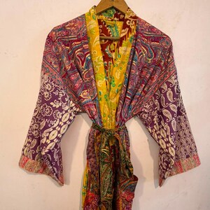 Może przedstawiać: Szlafrok typu kimono z długimi rękawami, wykonany z żywej patchworkowej tkaniny w wielu kolorach. Odzież posiada skomplikowane wzory paisley i kwiatowe w odcieniach ż&oacute;łci, fioletu, czerwieni i zieleni, wraz z pasującym paskiem do wiązania w talii.