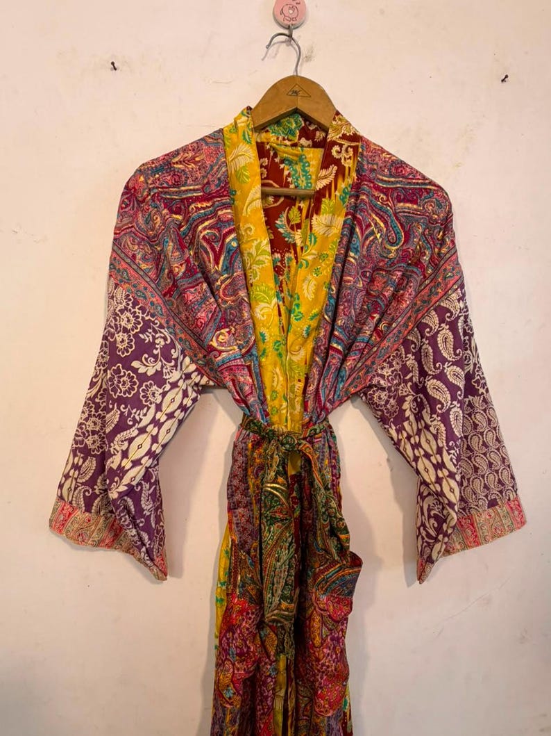 Może przedstawiać: Szlafrok typu kimono z długimi rękawami, wykonany z żywej patchworkowej tkaniny w wielu kolorach. Odzież posiada skomplikowane wzory paisley i kwiatowe w odcieniach ż&oacute;łci, fioletu, czerwieni i zieleni, wraz z pasującym paskiem do wiązania w talii.