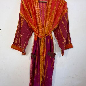 May include: A long-sleeved robe featuring a vibrant tie-dye pattern in shades of magenta and orange. The garment includes a matching tie belt at the waist, wide sleeves, and two front pockets, all displayed on a wooden hanger against a plain wall.