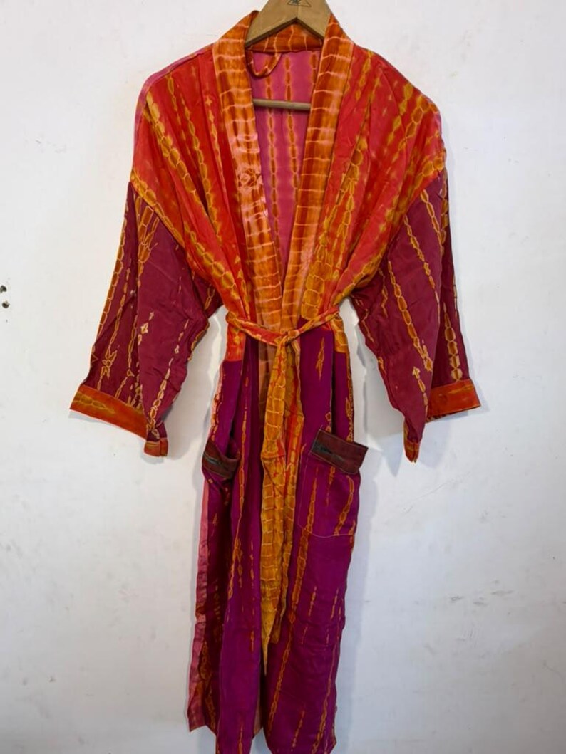 May include: A long-sleeved robe featuring a vibrant tie-dye pattern in shades of magenta and orange. The garment includes a matching tie belt at the waist, wide sleeves, and two front pockets, all displayed on a wooden hanger against a plain wall.