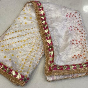 May include: White textile with yellow and orange dot patterns. The fabric has a decorative border with gold, pink, and white accents. The textile is likely a scarf or shawl, with a textured appearance.
