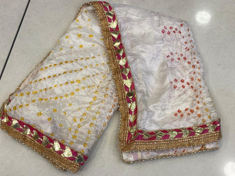 May include: White textile with yellow and orange dot patterns. The fabric has a decorative border with gold, pink, and white accents. The textile is likely a scarf or shawl, with a textured appearance.