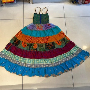 Könnte beinhalten: Ein bodenlanges Kleid mit gesmoktem Oberteil, dünnen Trägern und einem mehrstufigen Rock. Der Rock besteht aus horizontalen Bahnen in verschiedenen Farben und Mustern, darunter leuchtendes Blau, Orange, Blumenprints und ein Karomuster.