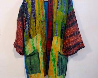 May include: A kimono-style jacket featuring a patchwork design with vibrant tie-dye patterns in yellow, green, blue, and deep red. The garment has wide sleeves, a solid blue inner lining, and two large front pockets, all displayed on a wooden hanger.