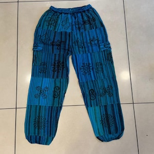 Puede incluir: Pantalones holgados con un patrón de rayas verticales en varios tonos de azul, incluyendo turquesa, marino y azul cielo. La tela está decorada con símbolos negros impresos, incluyendo soles y formas geométricas, y tiene una cintura elástica.