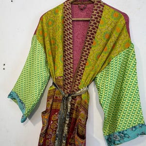 May include: A colorful, patterned robe with long sleeves and a matching belt. The robe features a mix of green, yellow, and red fabrics with floral and geometric designs. The sleeves have a green pattern with a blue floral trim. The robe is hanging on a wooden hanger.