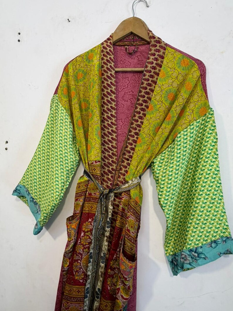 May include: A colorful, patterned robe with long sleeves and a matching belt. The robe features a mix of green, yellow, and red fabrics with floral and geometric designs. The sleeves have a green pattern with a blue floral trim. The robe is hanging on a wooden hanger.