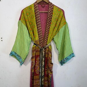 May include: A colorful, patterned robe with long sleeves and a tie waist. The robe features a mix of floral and geometric designs in shades of green, red, and purple. It has pockets and is hanging on a wooden hanger.