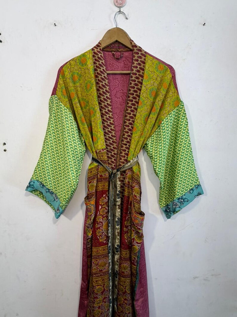 May include: A colorful, patterned robe with long sleeves and a tie waist. The robe features a mix of floral and geometric designs in shades of green, red, and purple. It has pockets and is hanging on a wooden hanger.