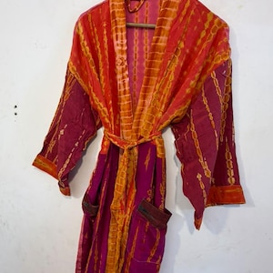 May include: A long-sleeved robe featuring a vibrant tie-dye pattern in shades of magenta, orange, and red. The garment includes a matching tie belt at the waist, two front pockets, and a shawl collar, all displayed on a wooden hanger.