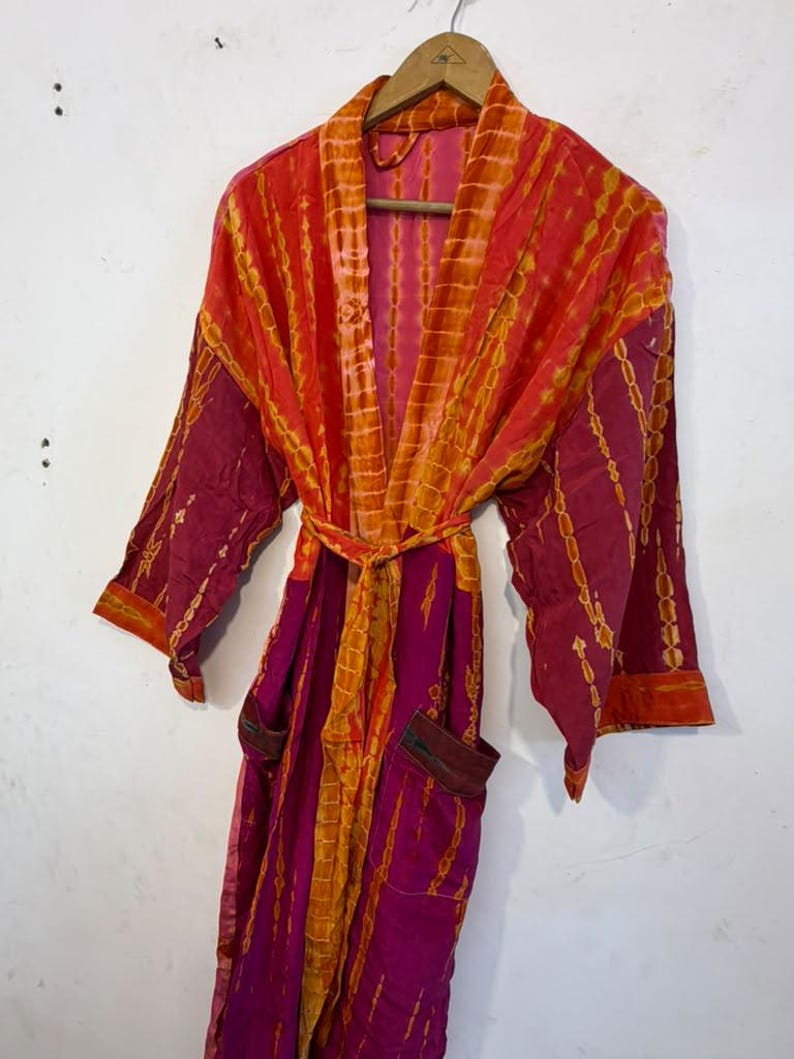 May include: A long-sleeved robe featuring a vibrant tie-dye pattern in shades of magenta, orange, and red. The garment includes a matching tie belt at the waist, two front pockets, and a shawl collar, all displayed on a wooden hanger.