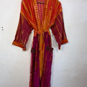 May include: A long-sleeved robe featuring a vibrant tie-dye pattern in shades of magenta and orange. The garment includes a matching tie belt at the waist, two front pockets, and a wide collar, all displayed hanging on a wooden hanger.
