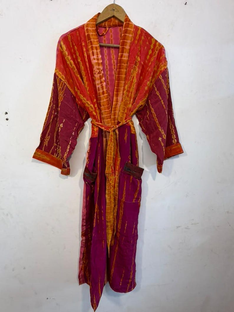 May include: A long-sleeved robe featuring a vibrant tie-dye pattern in shades of magenta and orange. The garment includes a matching tie belt at the waist, two front pockets, and a wide collar, all displayed hanging on a wooden hanger.