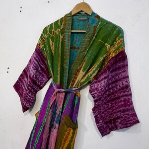 May include: A tie-dye patterned robe in shades of green, purple, and blue hangs on a wooden hanger. The robe features long sleeves, a waist tie, and two front pockets. The fabric has a textured appearance.