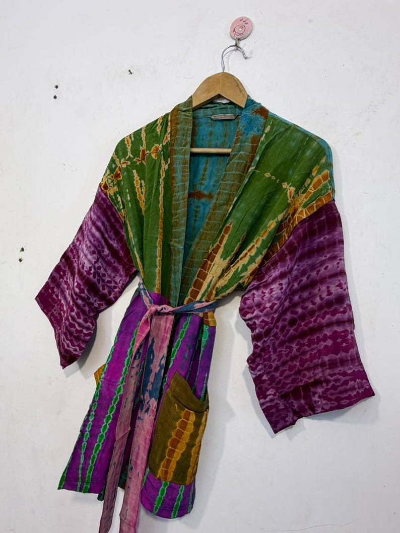 May include: A tie-dye patterned robe in shades of green, purple, and blue hangs on a wooden hanger. The robe features long sleeves, a waist tie, and two front pockets. The fabric has a textured appearance.