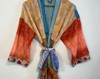 May include: A colorful, patterned robe with long sleeves, hanging on a wooden hanger. The robe features a mix of orange, red, yellow, and blue fabrics with floral and paisley designs. A matching belt is tied around the waist.