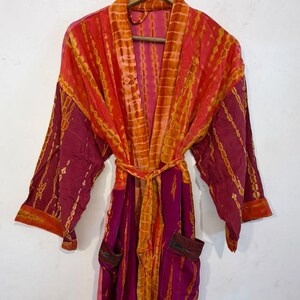 May include: A long-sleeved robe featuring a vibrant tie-dye pattern in shades of magenta, orange, and yellow. The garment includes a matching tie belt at the waist, two front pockets, and a wide collar, all displayed on a wooden hanger.