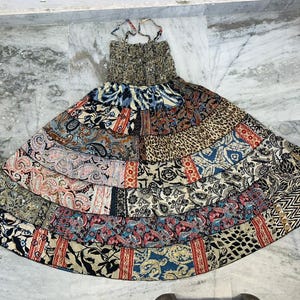 May include: A patchwork maxi dress with a tiered skirt. The dress features a variety of patterned fabrics in shades of blue, red, brown, and cream. The bodice is a textured, neutral color, and the straps are thin.