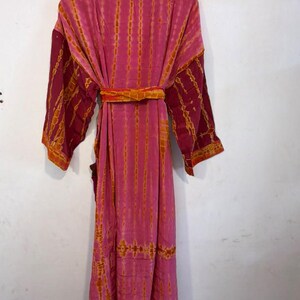 May include: A long-sleeved robe in shades of pink and orange featuring a vertical tie-dye pattern. The garment includes a matching fabric belt tied at the waist, a shawl collar, and wide sleeves, presented in a full-length design.