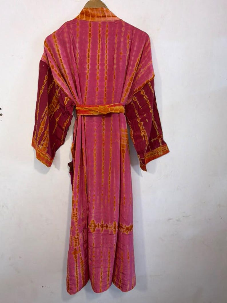 May include: A long-sleeved robe in shades of pink and orange featuring a vertical tie-dye pattern. The garment includes a matching fabric belt tied at the waist, a shawl collar, and wide sleeves, presented in a full-length design.