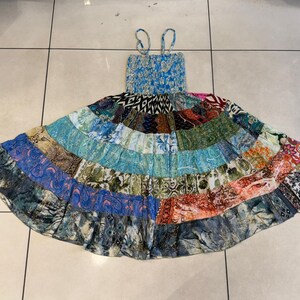 Puede incluir: Un vestido patchwork con corpi&ntilde;o azul y tirantes finos. La falda presenta una variedad de paneles de tela estampada en tonos azules, verdes, naranjas y rojos. El vestido est&aacute; tendido sobre una superficie embaldosada.