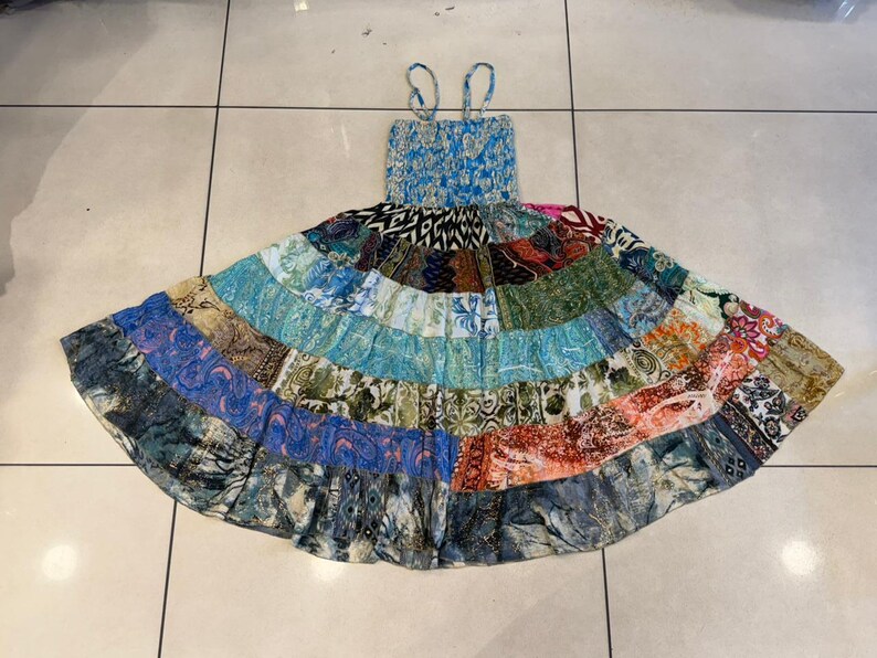 Puede incluir: Un vestido patchwork con corpi&ntilde;o azul y tirantes finos. La falda presenta una variedad de paneles de tela estampada en tonos azules, verdes, naranjas y rojos. El vestido est&aacute; tendido sobre una superficie embaldosada.