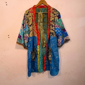 May include: A kimono-style robe featuring a vibrant patchwork of blue, red, green, and gold fabrics. The garment displays intricate paisley and floral patterns throughout, with a bright red trim along the front opening and two front pockets.