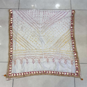 May include: White scarf with a geometric pattern in yellow and pink dots. The scarf has a decorative border with gold and red accents. The scarf is folded and displayed on a light-colored surface.