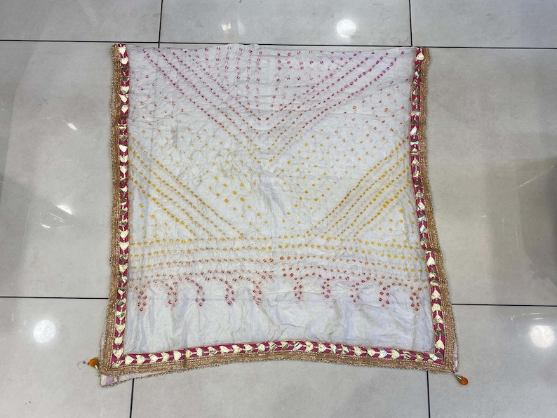 May include: White scarf with a geometric pattern in yellow and pink dots. The scarf has a decorative border with gold and red accents. The scarf is folded and displayed on a light-colored surface.