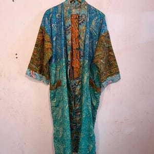 Może przedstawiać: Szlafrok typu kimono z długimi rękawami, ozdobiony żywą mieszanką wzorów paisley w odcieniach morskiej zieleni, błękitu i pomarańczu. Ubiór ma otwarty przód z kontrastującym pomarańczowo-niebieskim wykończeniem wzdłuż kołnierza oraz dwie kieszenie boczne.
