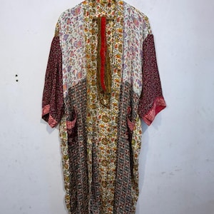 Può includere: Una lunga vestaglia kimono aperta sul davanti con un design patchwork. La vestaglia presenta motivi floreali e geometrici nei toni del rosso, bianco, giallo e marrone. Ha maniche larghe e due tasche anteriori, appesa a un appendiabiti di legno.