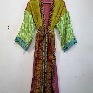 May include: A long, patchwork robe with a variety of patterns and colors, including green, red, and pink. The robe has long sleeves and a belt. The robe is hanging on a wooden hanger.