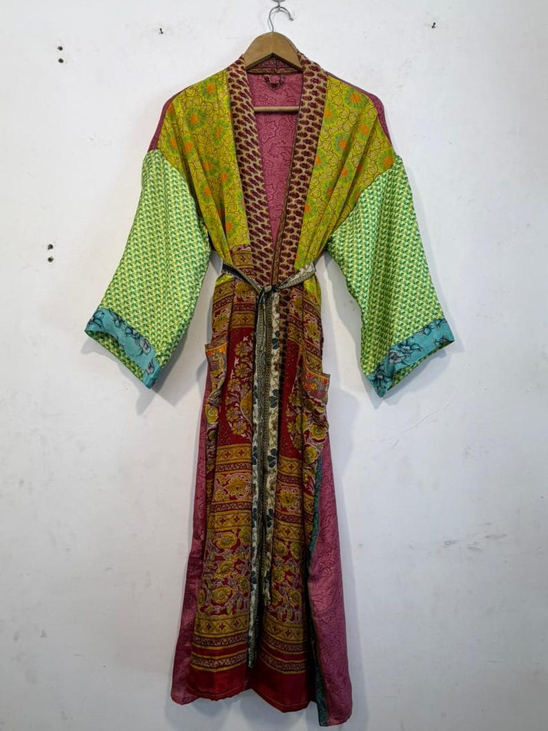 May include: A long, patchwork robe with a variety of patterns and colors, including green, red, and pink. The robe has long sleeves and a belt. The robe is hanging on a wooden hanger.