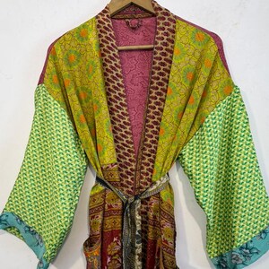 May include: A colorful, patterned robe with a belt, hanging on a wooden hanger. The robe features a mix of green, yellow, burgundy, and pink fabrics. The sleeves are a bright green with a geometric pattern, and the edges are trimmed with blue floral fabric.