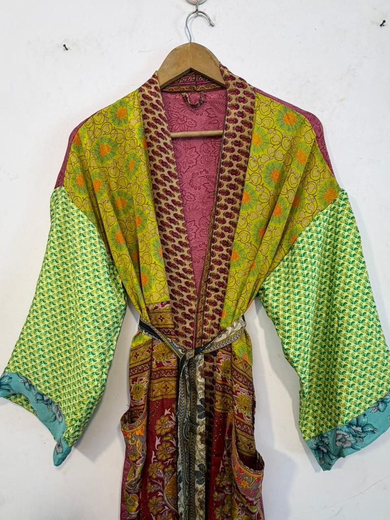 May include: A colorful, patterned robe with a belt, hanging on a wooden hanger. The robe features a mix of green, yellow, burgundy, and pink fabrics. The sleeves are a bright green with a geometric pattern, and the edges are trimmed with blue floral fabric.
