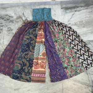 Puede incluir: Un pantalón palazzo patchwork con cintura elástica turquesa. El pantalón de pernera ancha presenta paneles con estampados de cachemir y geométricos en tonos azules, morados, rosas y verdes. El pantalón está extendido sobre una superficie de mármol.