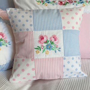 Könnte beinhalten: Quadratisches Patchwork-Kissen mit floraler Stickerei. Das Kissen hat eine Mischung aus weißen Punkten, rosa Streifen und blauen Streifen. Die gestickten Blumen sind rosa und blau mit grünen Blättern.