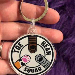 May include: A white round keychain with black text that reads "Tof Bean Squad" and three paw prints in the center. The paw prints are brown, pink, and gray.