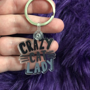 May include: A clear acrylic keychain with the words "CRAZY CAT LADY" in black and gray lettering. The keychain has a silver metal ring for attaching keys. The background is a fuzzy purple material.