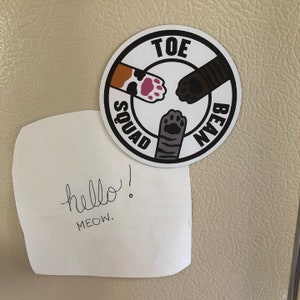 May include: A white round sticker with black text and paw prints. The text reads "Toe Bean Squad". The sticker is on a beige surface with a white note that says "hello! MEOW."