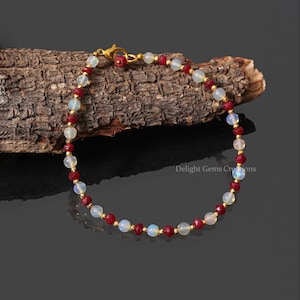 Könnte beinhalten: Ein zartes Armband aus abwechselnd weißen Opal- und roten Edelsteinperlen. Das Armband ist mit einem goldenen Verschluss versehen.