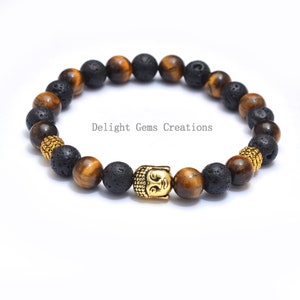 Puede incluir: Una pulsera de cuentas con una cabeza de Buda dorada, ojo de tigre, roca volcánica y cuentas doradas. La pulsera está ensartada en una banda elástica. El texto "Delight Gems Creations" es visible.