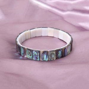 Può includere: Un braccialetto elastico composto da segmenti rettangolari. La superficie esterna presenta tessere in conchiglia di abalone iridescente con motivi vorticosi verdi, blu e viola, mentre il lato interno è costituito da tessere in conchiglia bianca liscia.