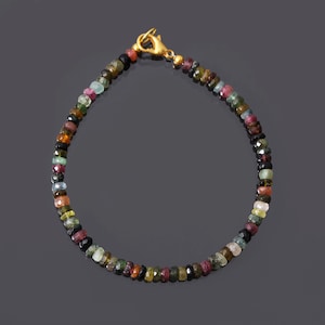 Puede incluir: Un brazalete de piedras preciosas multicolores con un cierre de oro. El brazalete está hecho de pequeñas cuentas redondas en tonos de verde, rosa, rojo, negro y azul.