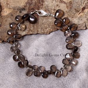 May include: A bracelet made of smoky quartz teardrop beads, featuring a silver clasp. The translucent brown beads are strung on a clear cord. The bracelet is displayed on a textured, brown surface.