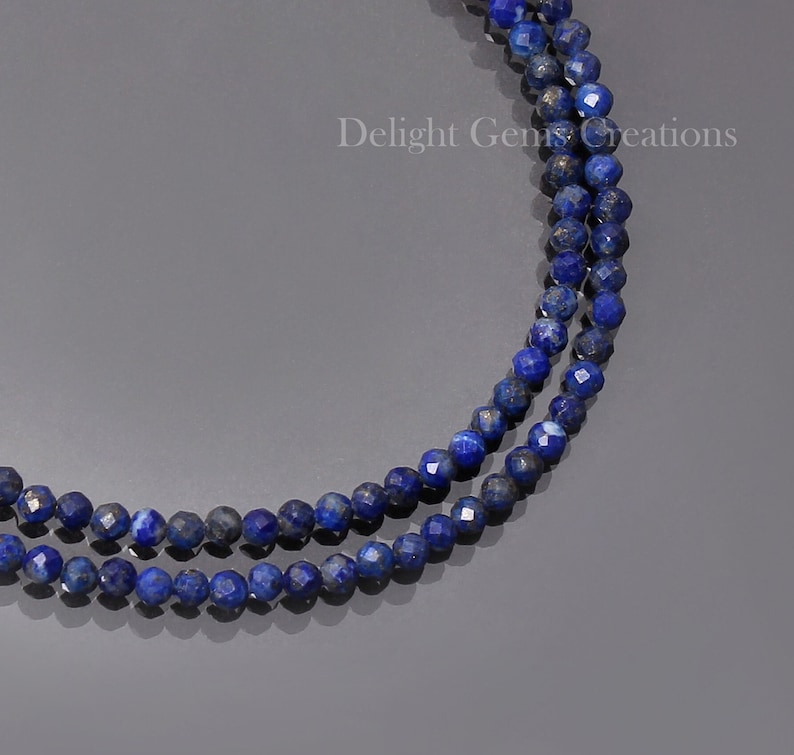 May include: A double strand necklace made of dark blue lapis lazuli beads. The beads are faceted and have a slightly metallic sheen. The necklace is on a grey background. Delight Gems Creations is printed on the background.