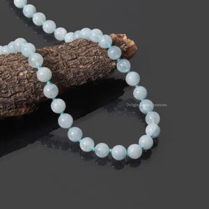 Op de afbeelding: Een lichtblauwe aquamarijn kralenketting. De ronde kralen zijn geregen op een bijpassend lichtblauw koord. De ketting is gedrapeerd over een stuk bruin hout, tegen een donkere achtergrond.