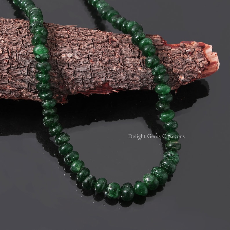 Puede incluir: Un collar de cuentas de piedras preciosas pulidas, de color verde oscuro. Las cuentas est&aacute;n ensartadas, creando una forma suave y redondeada. El collar se exhibe sobre un trozo de madera texturizada sobre un fondo oscuro. El texto "Delight Gems Creations" es visible.