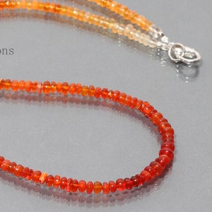 May include: A necklace made of small, round, orange opal beads. The beads are graduated in size, with the larger beads at the bottom of the necklace. The necklace is strung on a silver chain.