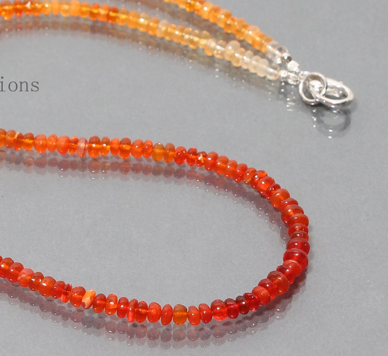 May include: A necklace made of small, round, orange opal beads. The beads are graduated in size, with the larger beads at the bottom of the necklace. The necklace is strung on a silver chain.